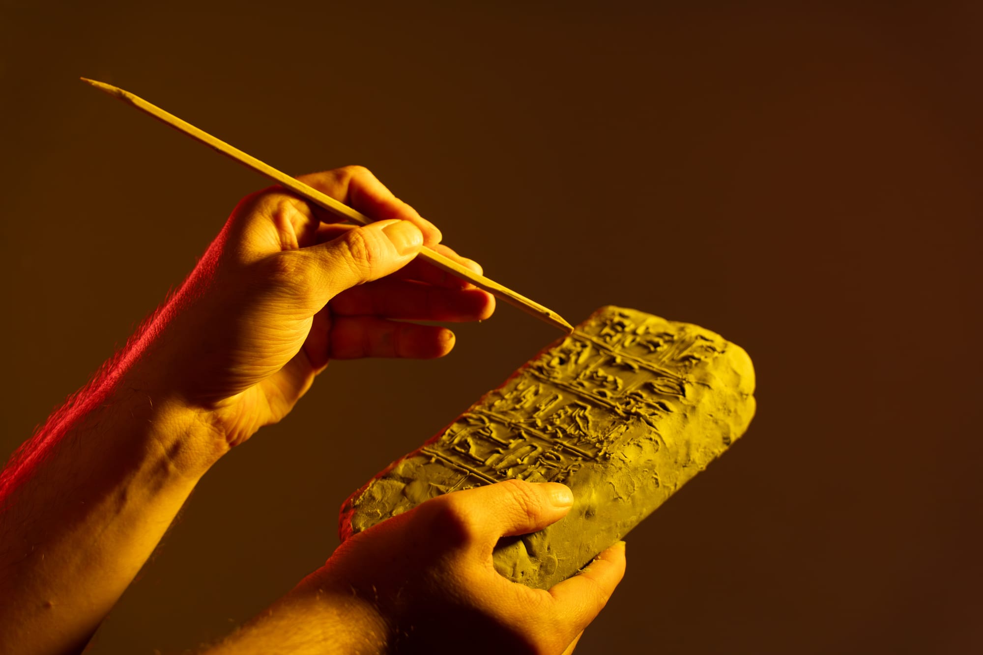 Hands holding a clay tablet inscribed with cuneiform script while using a stylus to trace the ancient wedge-shaped characters
