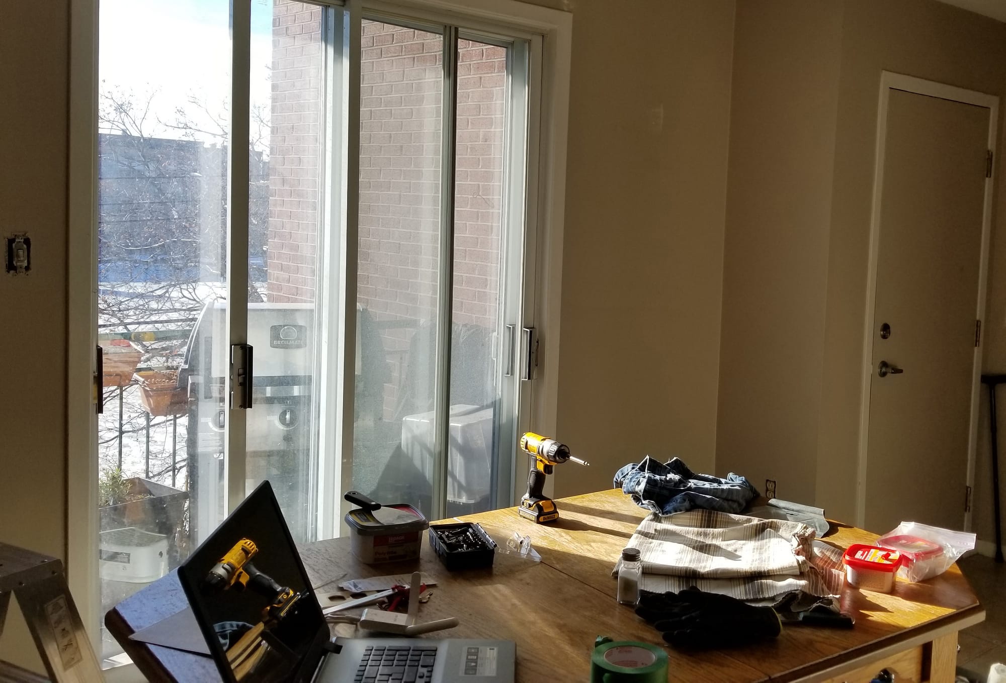 Sunlit dining area with a cluttered table, tools, and a laptop, representing a messy, unfinished living space.